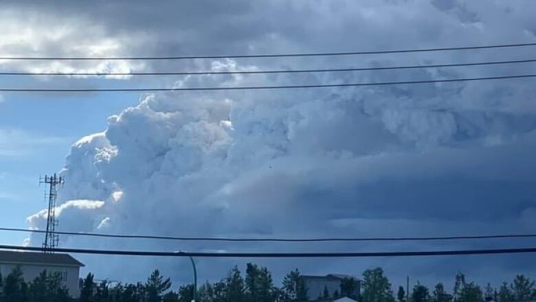 Cumulonimbus clouds with rings of smoke visible at the bottom pile high into the air over trees and houses.