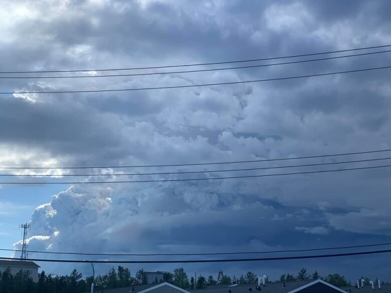 Cumulonimbus clouds with rings of smoke visible at the bottom pile high into the air over trees and houses.