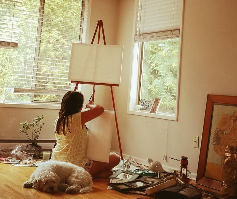 A young girl sits with her back to the camera, in front of an art easel in the corner of a room.