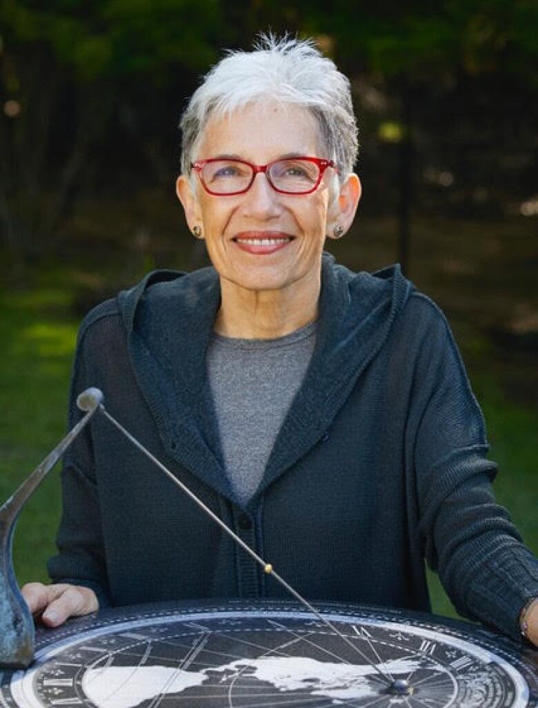 Dava Sobel poses for the camera in front of a sundial. She has white hair, cut short, and is wearing red-rimmed glasses.