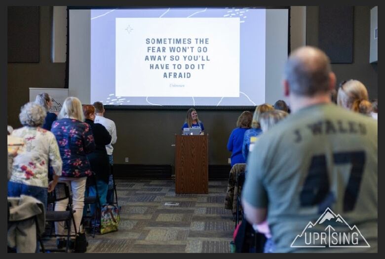 A woman pictured at a pulpit in a conference hall with a projector screen behind her, displaying the words, 'Sometimes the fear won't go away so you'll have to do it afraid.'