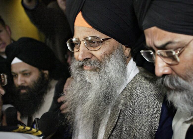 A man with a long grey beard and glasses is flanked by other men at a press scrum. He is wearing a black turban and a grey suit.