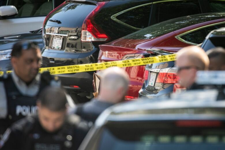 A number of police officers are seen near yellow crime tape and a red sedan.