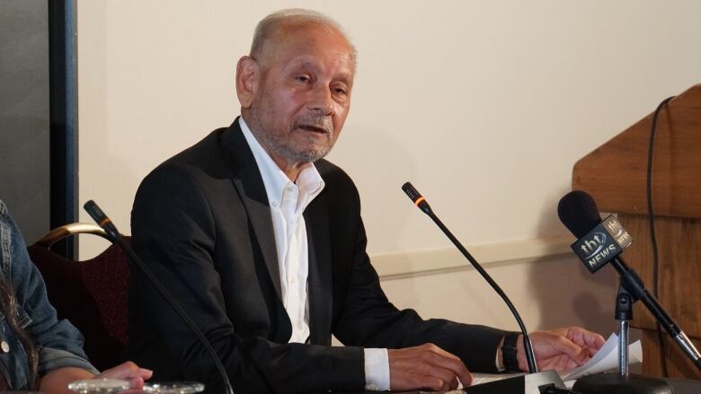 An older man wears a suit and sits at a table speaking into a microphone.