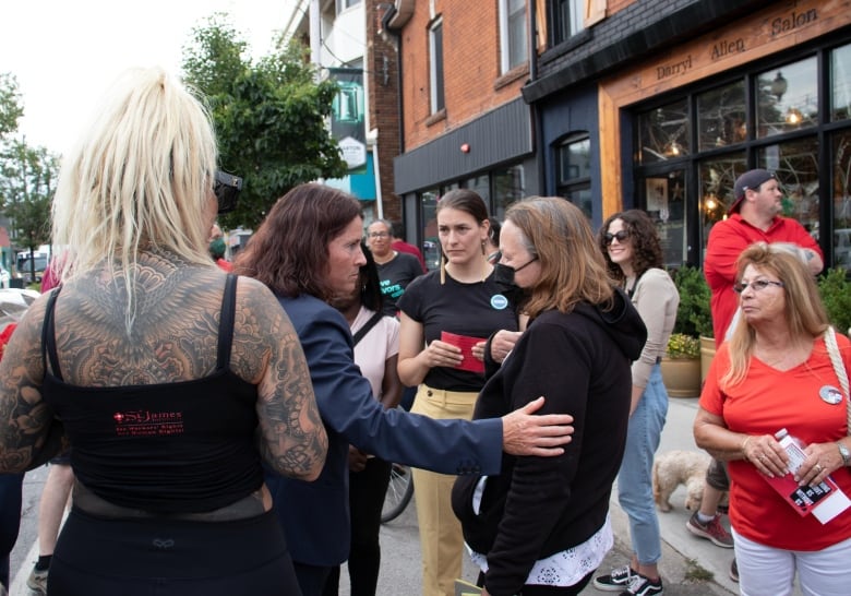 A police detective has her hand on the arm of a distraught woman. They are surrounded by a crowd of supporters who also look concerned. 