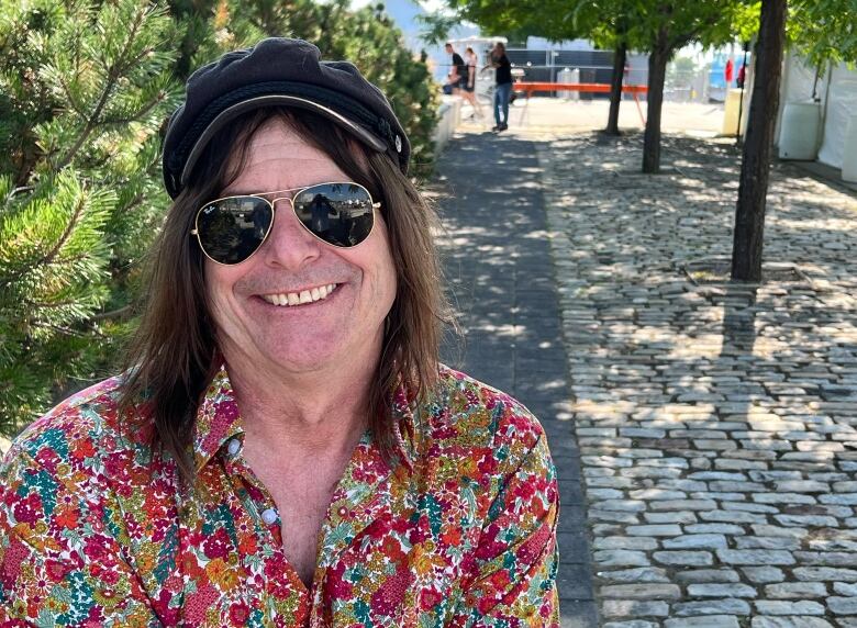 A man wearing sunglasses, a cap and a brightly-coloured shirt covered in a flower design smiles at the camera.