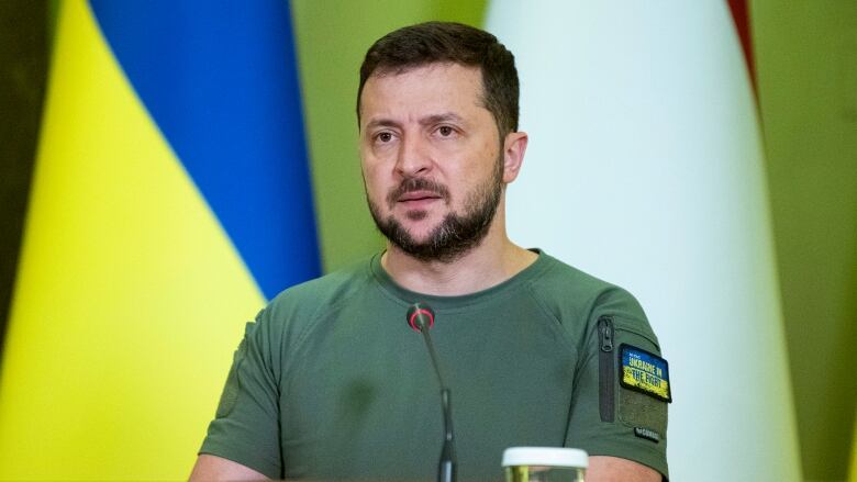 A man in a green military tshirt is shown speaking at a podium in front of a Ukrainian flag.