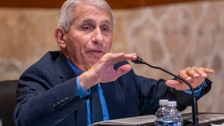 A grey-haired man in a suit gestures while speaking into a microphone.