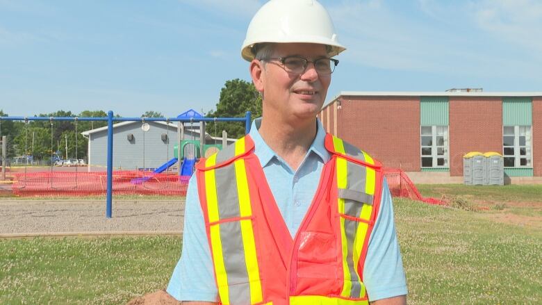 Man wearing hard hat, glasses and a safety vest stands outside a school.