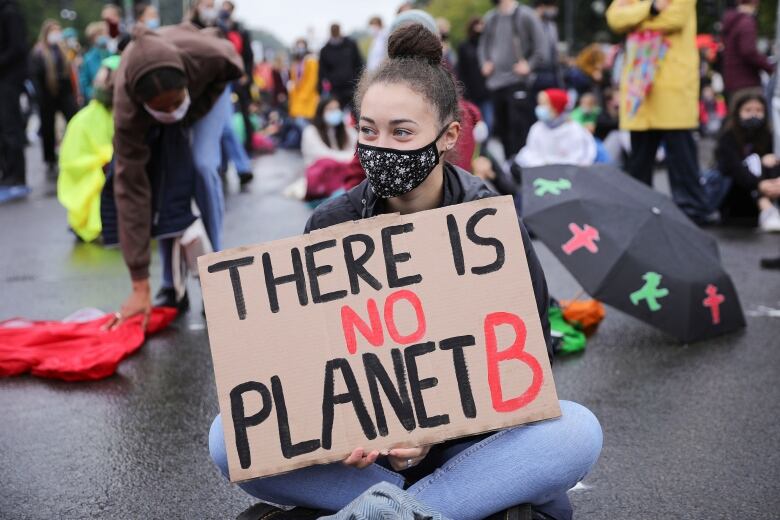 A young woman sits on the protest at a protest. She is holding a sign that says 'There is no Planet B.'