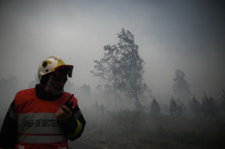 An emergency worker wearing a helmet and orange safety vest talks into a walkie-talkie while walking through a smoke-filled field.