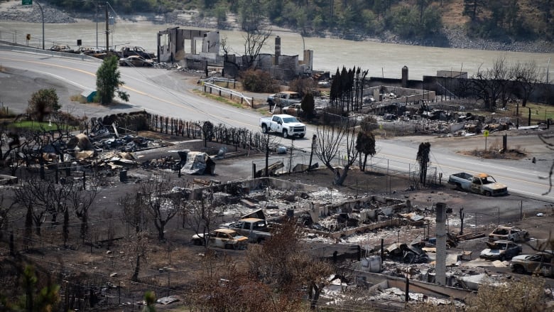 A white police truck drives down a road past shells of burned-out vehicles and structures destroyed by a wildfire. 