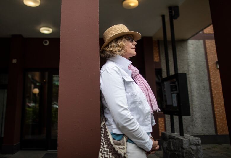 Susan MacRae, a blonde woman wearing a wide-brimmed hat, is pictured in the West End in Vancouver on Monday, July 18, 2022. MacRae is shown in profile, leaning against a brown painted post in front of an apartment building, and is dressed in light pants, a white denim jacket, turquoise shirt and pink scarf. 