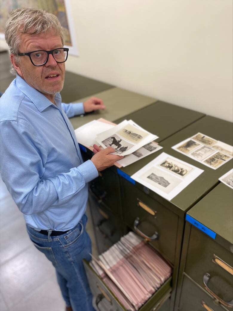 A grey-haired man in shirt and jeans stands by an open filing cabinet, examining archival photos.