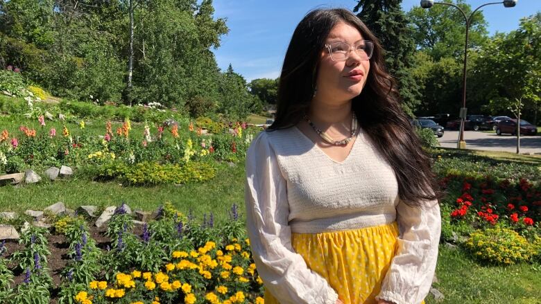 A young woman stands in a park with flowers in the background. 