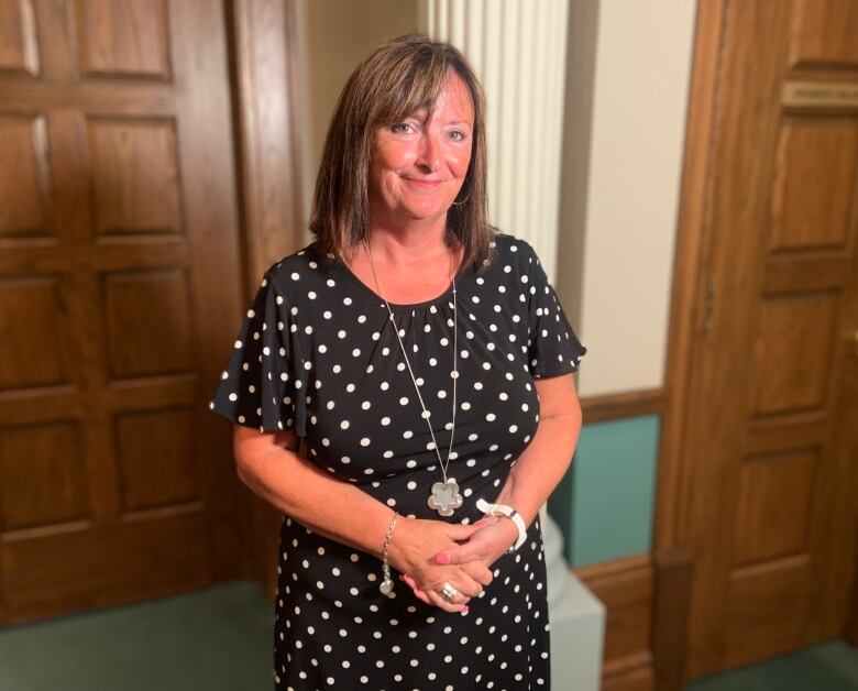 A person with long brown hair and a black and white polka dot dress smiles at a camera, standing in front of a column outside the Newfoundland and Labrador House of Assembly.