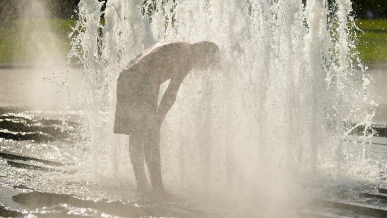 A person bends over and stands in the middle of a fountain to cool down.