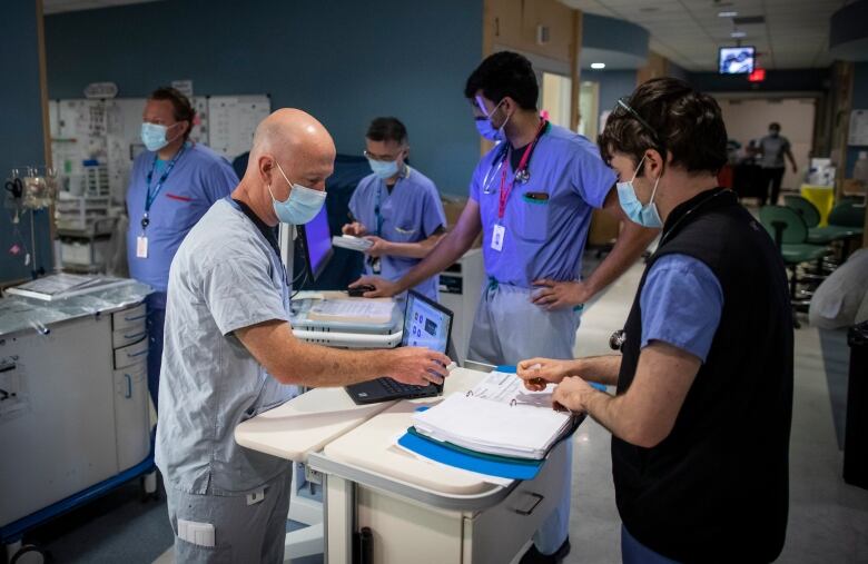 Five health-care professionals wearing blue scrubs and medical masks look at electronic and paper documents in a hospital intensive care unit.