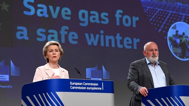 A woman and a man are shown at podiums, in front of a screen that says 'Save gas for a safe winter.'