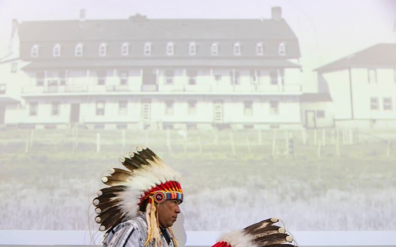 A First Nations man in a headdress walks past a projected image of an old, three-storey building.