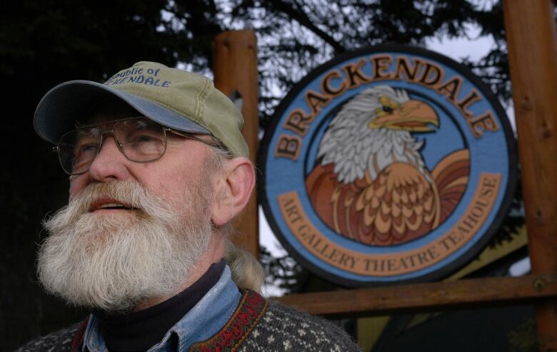 An old man with a white beard poses in front of an eagle symbol.