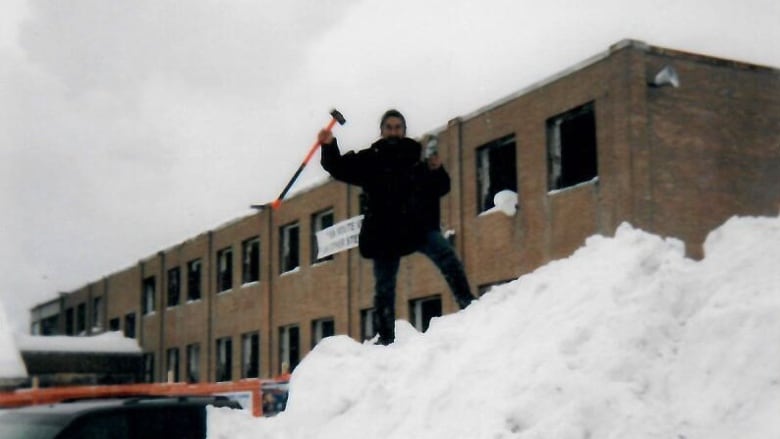 A man stands on a pile of snow outside a dilapidated building.