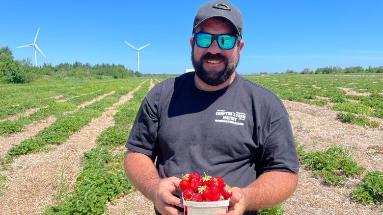 Man wearing baseball hat and sunglasses holding basket of strawberries in a strawberry field.