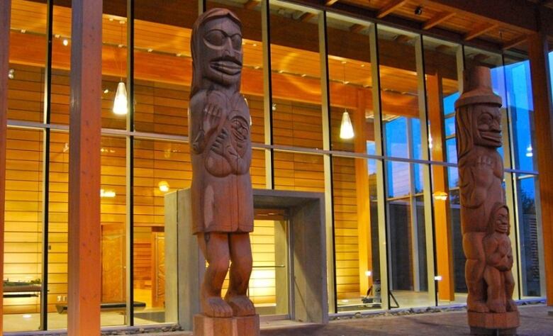 Two Indigenous artifacts stand in front of a glass building.