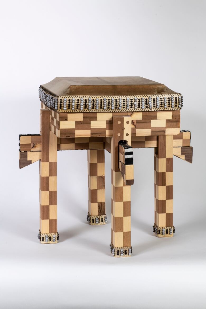 Photo of a wooden drum in an all-white space. It is in the form of a bench and the wood is interlocked in a checkerboard pattern of two shades of wood. The rim of the drum and the feet are adorned with black and white beads.