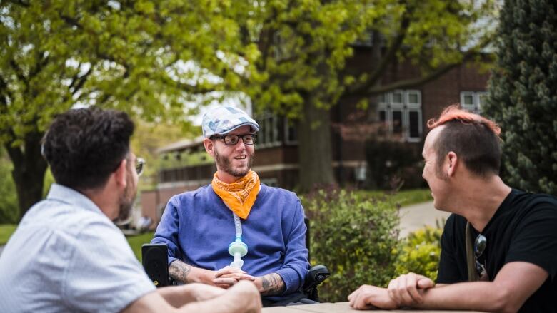 Three people sit at a picnic table. Two are facing away from the camera. The person in the centre is facing the camera, and has glasses, a beard and a hat on.