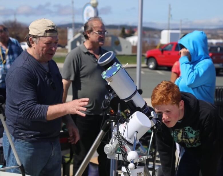 A boy with red hair leans to looking into a telescope, while an older man wearing a ball cap gives him direction. 