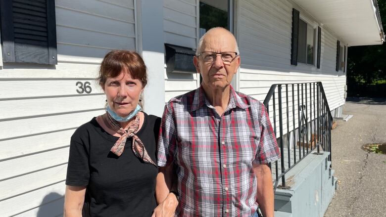 Elderly couple stand in front of a building, arms linked.