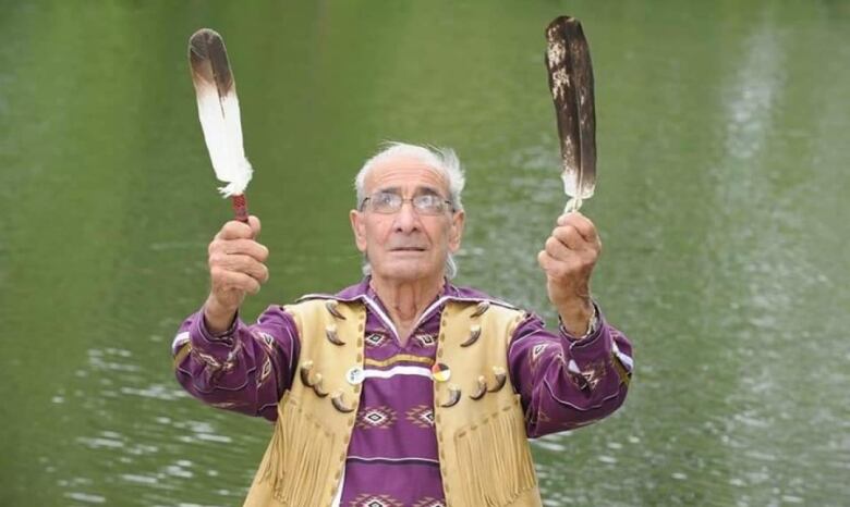 An older man holds up two feathers by a pond.