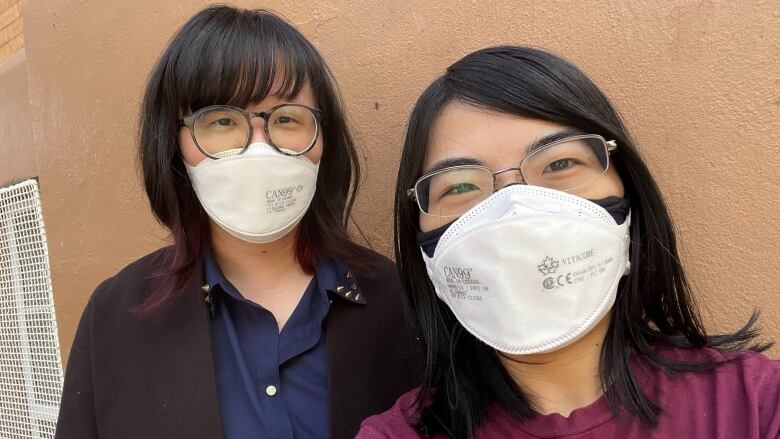 Two people wearing white N95 masks look at the camera. They are both wearing glasses and appear to be smiling behind the masks.