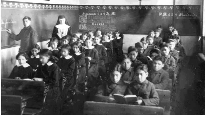 A black-and-white historical photo shows a nun standing in a room of Indigenous boys and girls seated at desks. One student stands beside the nun, writing on a chalkboard.