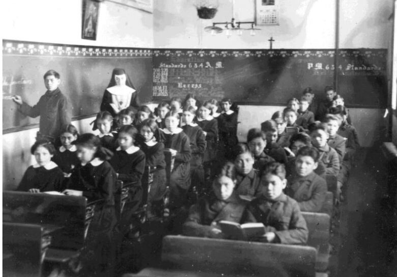 A black-and-white historical photo shows a nun standing in a room of Indigenous boys and girls seated at desks. One student stands beside the nun, writing on a chalkboard.