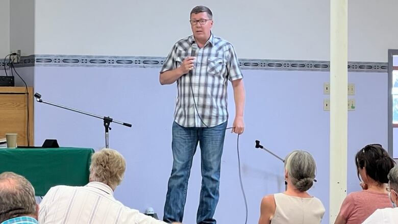 A man in blue jeans and a checkered shirt stands on a stage holding a microphone, speaking to a group of seated people.