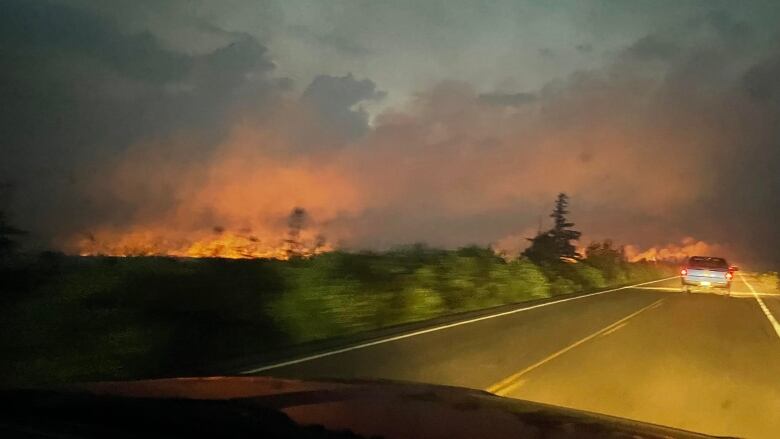 A photograph taken from the front window of a pick up truck shows flames along the left side of the highway and another truck ahead on the road.