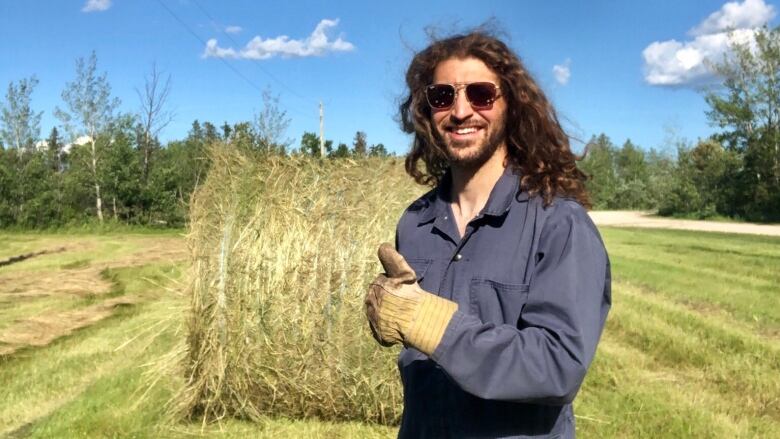 A man with long dark hair and a beard stands in a field in front of a hay bale. He wears sunglasses, blue coveralls and a leather glove, and gives a thumbs up to the camera. 