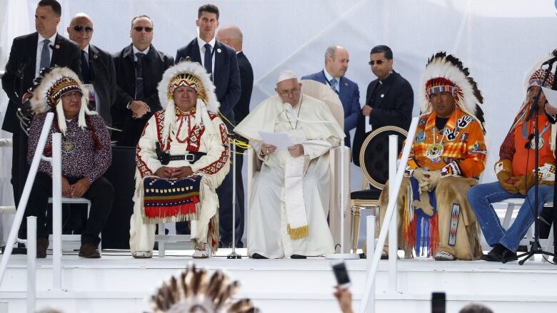 Pope Francis sits on a stage surrounded by First Nations chiefs in headdresses.