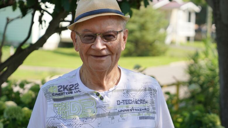 An elderly man stands outside in the sun, smiling. 