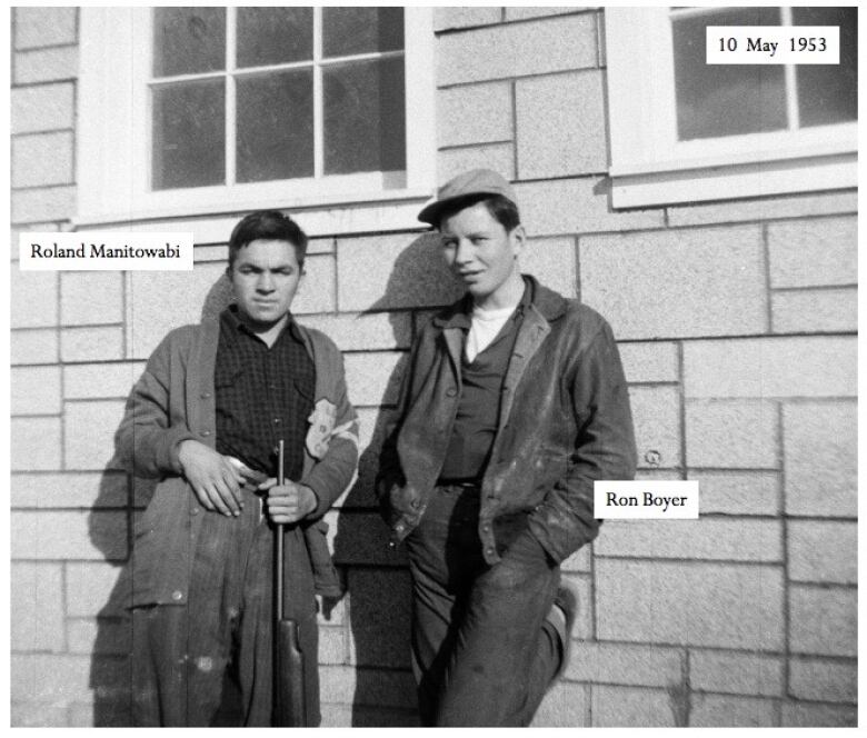 Two young Mohawk teens stand outside a residential school. The name Roland Manitowabi is written next next to the one on the left, Ron Boyer alongside the boy on the right. A date is noted at the top reading May 10, 1945.