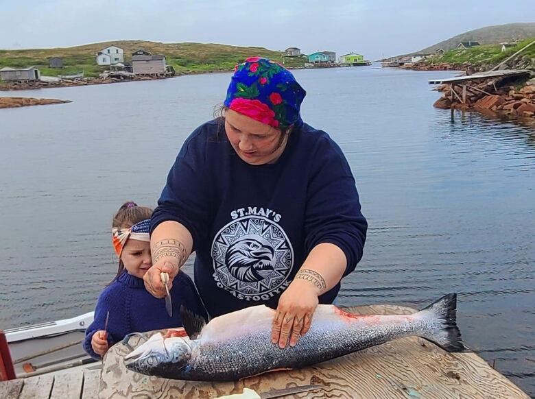 A woman uses a crescent-shaped knife to gut and clean a salmon on a wharf while a young girl looks on.