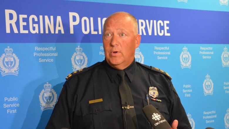 Regina Police Service Chief Evan Bray stands in front of a blue background emblazoned with the service's logo and motto. 