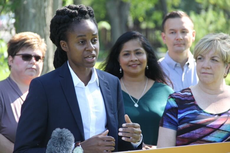 A person wearing a white shirt and dark blue blazer, surrounded by several other people, speaks at a podium in a park.