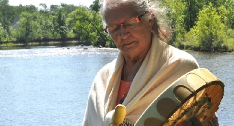 A woman holds a hand drum in front of a river.