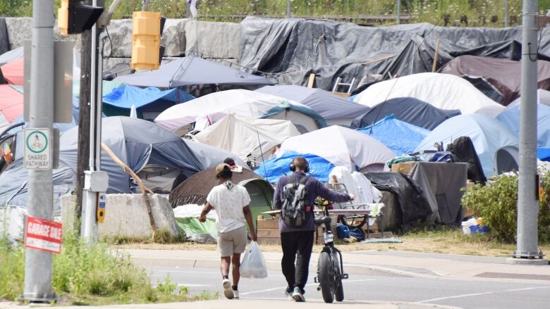 Two people, one walking beside a bicycle, cross at an intersection and walk towards multiple tents.