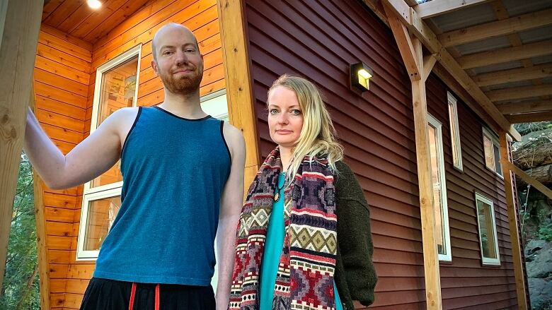 Bryce and Saige look at the camera from the porch of their tiny home. Bryce is wearing a blue sleeveless top, and Saige is wearing a long scarf atop a blue top.