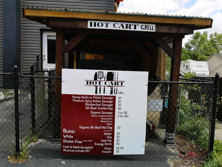 A wooden pergola is over a hot dog cart, which is behind a large sign that says 