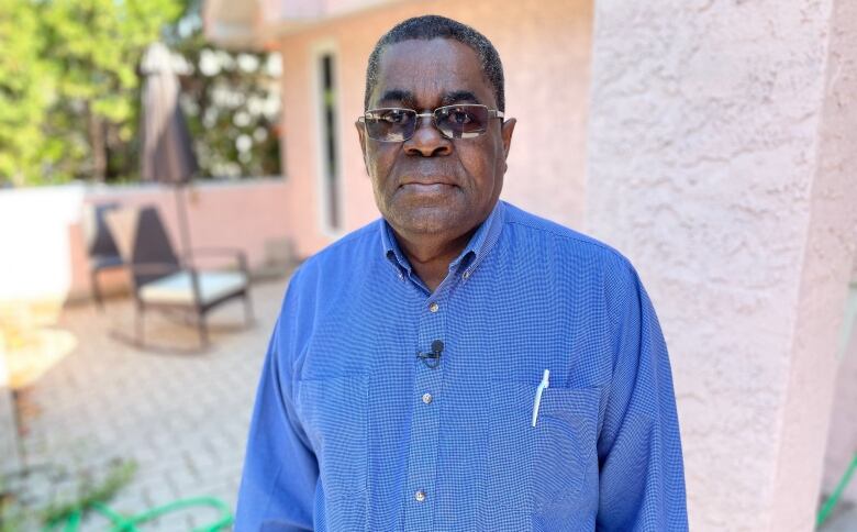 A man standing outside a home, wearing a blue shirt and sunglasses, looks at the camera.